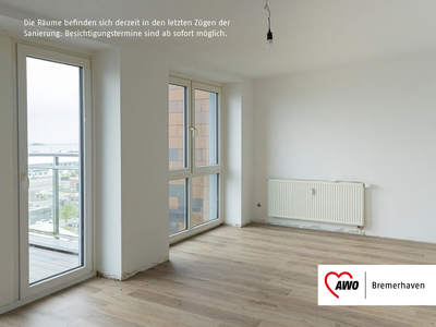 Wohnzimmer mit Ausblick | © AWO Bremerhaven Heller leerer Raum mit großen Fenstern, Holzboden, Heizkörper an der Wand und Zugang zu einem Balkon mit Glasgeländer. | © AWO Bremerhaven